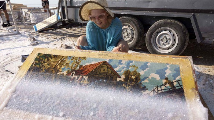 Potret Unik Karya Seni dari Kristal Garam di Laut Mati Multidisciplinary Israeli artist Sigalit Landau inspects one of her artworks, after being submerged in the salty Dead Sea water at the Ein Bokek resort, on August 2, 2022. - Israeli artist Sigalit Landau wades into the warm, briny waters of the Dead Sea to inspect her latest creations -- everyday objects coated in salt crystals that glisten in the bright morning sun. The lowest point on Earth is also Landau's studio where she submerges objects -- from a ballet dress to a lampshade's wire frame -- for weeks until they are magically transformed by ice-like layers of salt. (Photo by MENAHEM KAHANA / AFP) (Photo by MENAHEM KAHANA/AFP via Getty Images)