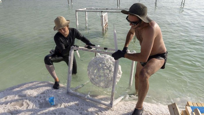 Potret Unik Karya Seni dari Kristal Garam di Laut Mati Multidisciplinary Israeli artist Sigalit Landau inspects one of her artworks, after being submerged in the salty Dead Sea water at the Ein Bokek resort, on August 2, 2022. - Israeli artist Sigalit Landau wades into the warm, briny waters of the Dead Sea to inspect her latest creations -- everyday objects coated in salt crystals that glisten in the bright morning sun. The lowest point on Earth is also Landau's studio where she submerges objects -- from a ballet dress to a lampshade's wire frame -- for weeks until they are magically transformed by ice-like layers of salt. (Photo by MENAHEM KAHANA / AFP) (Photo by MENAHEM KAHANA/AFP via Getty Images)