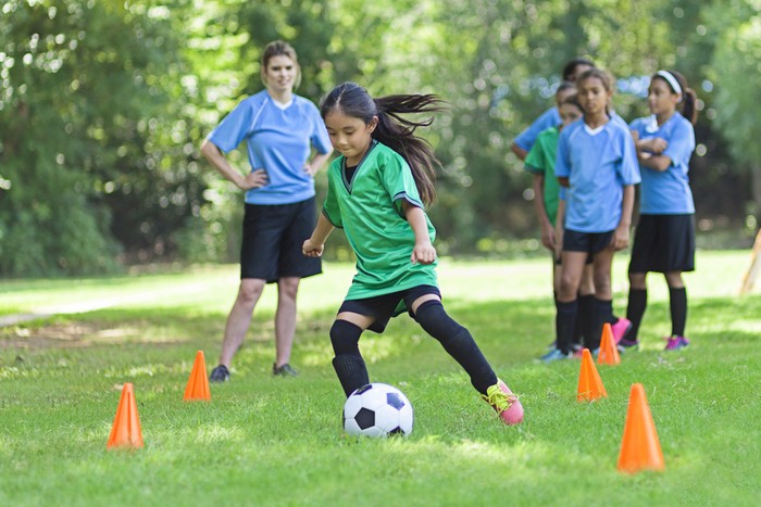Asian elementary age soccer athlete confidently kicks the ball around orange practice cones. She is wearing a green uniform. Players in blue uniforms are lined up behind her as well as her young adult Caucasian female coach.