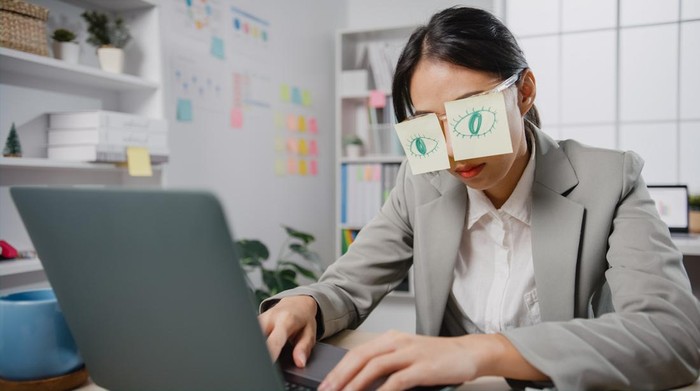 Tren baru quiet quitting Young Asian businesswoman sitting on desk overworked tired sleep over a laptop at office. Exhausted burnout lady with two post-it over her eyes, adhesive notes on face sleeping at workplace.