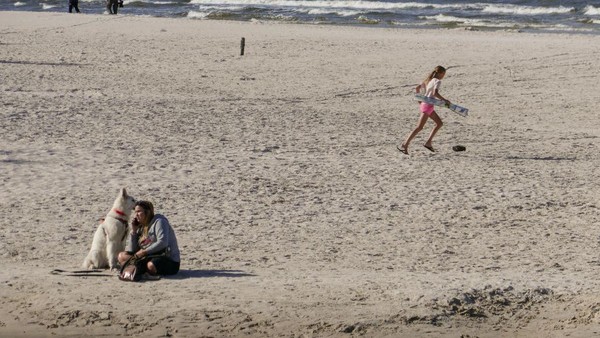 Seorang wanita bersama anjing peliharaannya duduk di pinggir pantai laut Baltik, Debki, Polandia sambil berjemur dan melihat aktifitas warga lainnya. Terlihat anak kecil yang asik bermain di belakangnya.