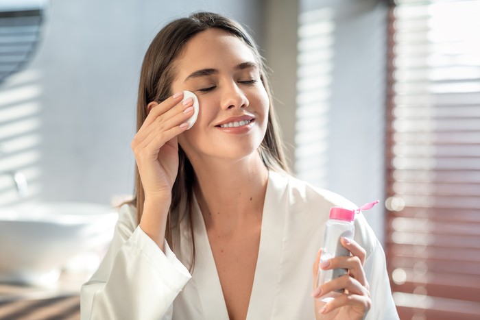 Ilustrasi Micellar Water Beautiful Smiling Woman Cleansing Skin With Micellar Water And Cotton Pad While Making Daily Beauty Routine In Bathroom, Attractive Young Female Enjoying Home Skincare Treatments, Copy Space
