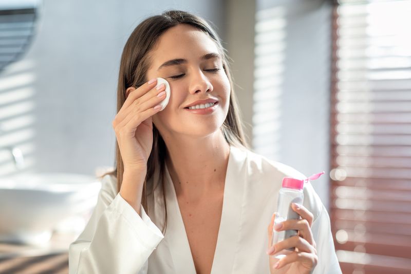 Beautiful Smiling Woman Cleansing Skin With Micellar Water And Cotton Pad While Making Daily Beauty Routine In Bathroom, Attractive Young Female Enjoying Home Skincare Treatments, Copy Space