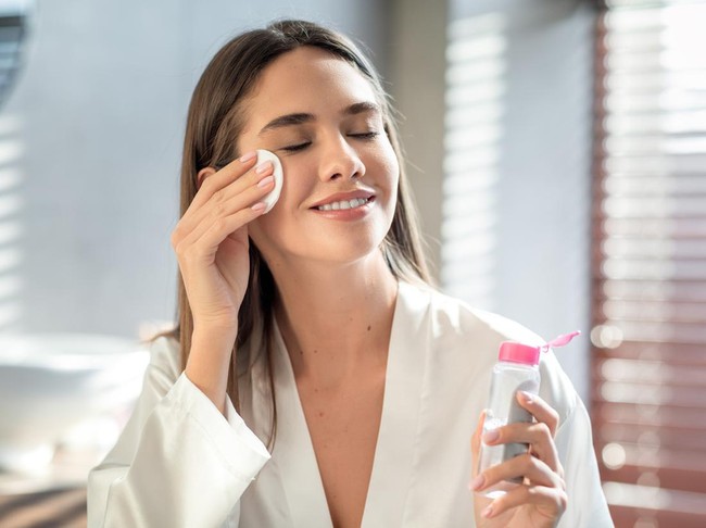 Beautiful Smiling Woman Cleansing Skin With Micellar Water And Cotton Pad While Making Daily Beauty Routine In Bathroom, Attractive Young Female Enjoying Home Skincare Treatments, Copy Space