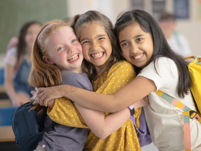 Three elementary school girls laugh and smile as the hug each other and look at the camera. They are wearing their backpacks and standing in a classroom.