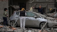 Warga membersihkan mobil di dekat bangunan tempat tinggal yang rusak akibat serangan militer Rusia. Mobil tersebut nampak rusak berat tertimpa puing apartemen.