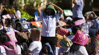 Seorang siswa berdiri di tengah teman-temannya yang sedang duduk. Para siswa tersebut juga diberikan permadani di kepalanya untuk perlindungan selama mengikuti latihan gempa.