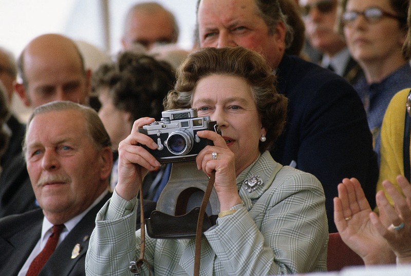 British Royal Queen Elizabeth II, wearing a headscarf and a waxed jacket, takes a picture with her Leica M6 camera at the Royal Windsor Horse Show, held at Windsor Home Park in Windsor, Berkshire, England, 12th May 1989. (Photo by Tim Graham Photo Library via Getty Images)