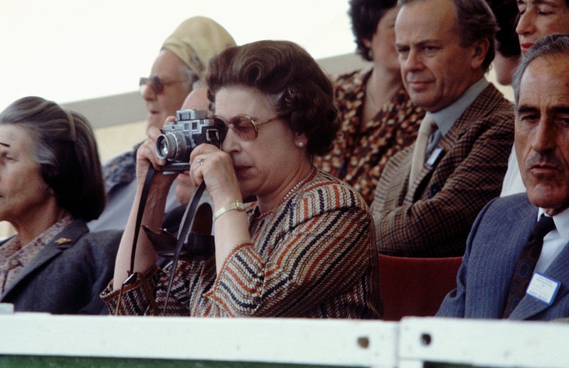 British Royal Queen Elizabeth II, wearing a headscarf and a waxed jacket, takes a picture with her Leica M6 camera at the Royal Windsor Horse Show, held at Windsor Home Park in Windsor, Berkshire, England, 12th May 1989. (Photo by Tim Graham Photo Library via Getty Images)