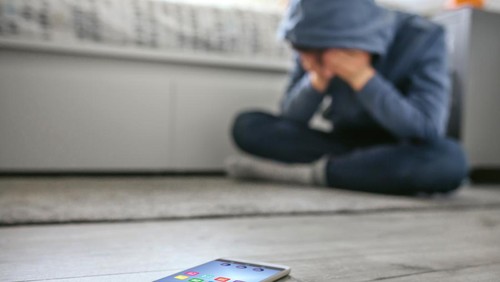 Unrecognizable teenager boy crying desperate for bullying with mobile lying on the floor in foreground. Selective focus on mobile in foreground