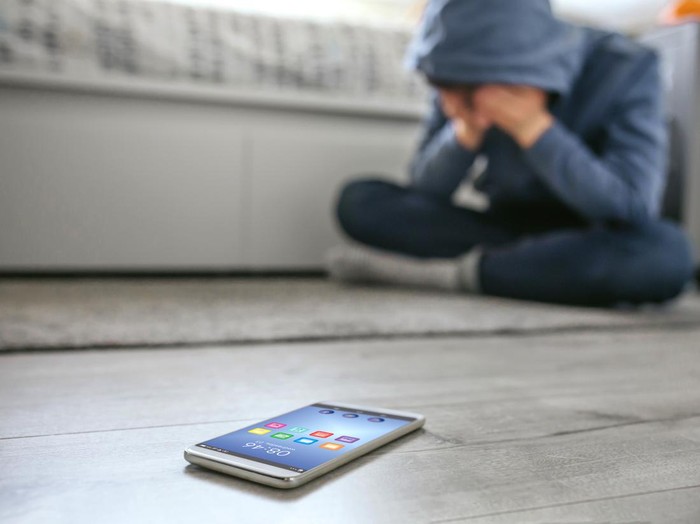 Unrecognizable teenager boy crying desperate for bullying with mobile lying on the floor in foreground. Selective focus on mobile in foreground