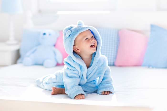Ilustrasi Cara Agar Rambut Bayi Tumbuh Lebat Cute happy laughing baby boy in soft bathrobe after bath playing on white bed with blue and pink pillows in sunny kids room. Child in clean and dry towel. Wash, infant hygiene, health and skin care.