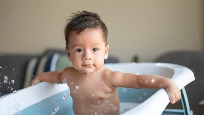 Adorable six moths old mixed race baby boy taking a bath in a small baby tub in the living room splashing water excitedly