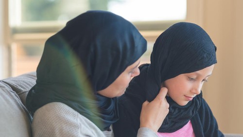 An Arab-American woman reads and snuggles with her elementary age daughter in her lap. They are both wearing traditional headscarves.
