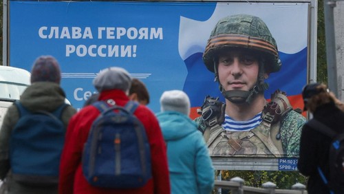 People gather at a tram stop in front of a board displaying a portrait of Russian service member Sergei Tserkovniy in Saint Petersburg, Russia September 21, 2022. A slogan on the board reads: Glory to heroes of Russia!  REUTERS/Anton Vaganov