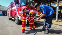 Dua orang anak TK tersebut mengenakan pakaian pemadam kebakaran lengkap. Seorang petugas terlihat sedang memberikan arahan kepada mereka.