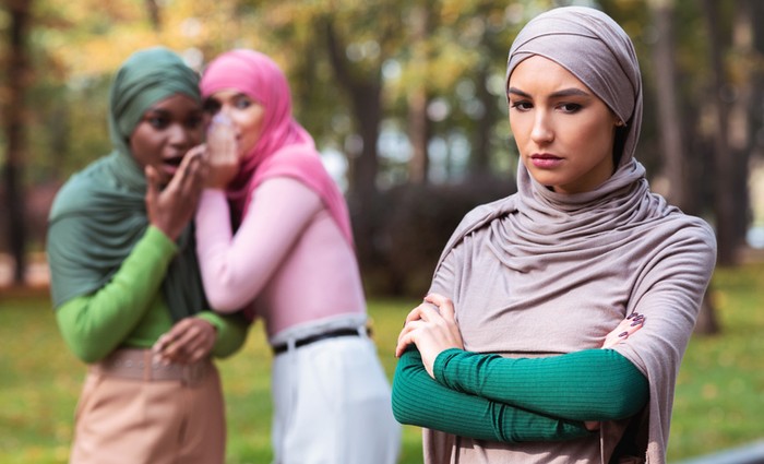 Bullying. Fake Friends Talking About Unhappy Muslim Woman Standing Alone In Park Outdoor. Female Friendship Problem, Outcast Person Concept. Selective Focus