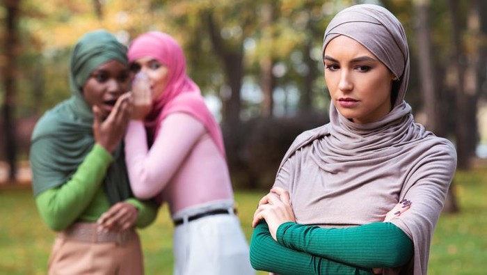 Bullying. Fake Friends Talking About Unhappy Muslim Woman Standing Alone In Park Outdoor. Female Friendship Problem, Outcast Person Concept. Selective Focus