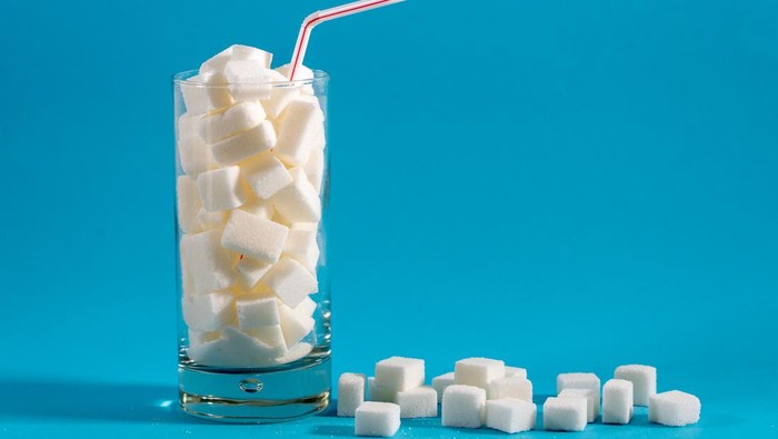 Glass with red stripes straw filled with sugar cubes on blue background. Junk food, unhealthy diet, sugar on refreshing drinks, nutrition concept.