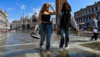 Turis berjalan melintasi alun-alun St. Mark yang banjir dengan menenteng alas kakinya di Venesia.