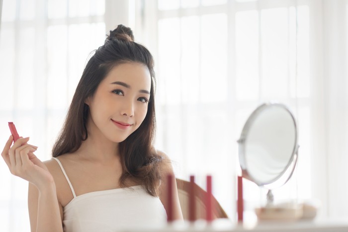 Beautiful Asian Female in white vest holding a red or pink lipstick and looking at camera. Happy woman applying makeup. Blogger, Cosmetics, Hairstyle.