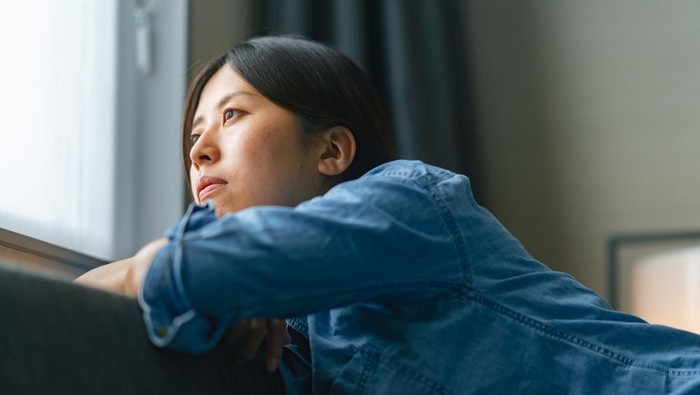 Ilustrasi Wanita Sedih dan Termenung A young woman is sitting on a sofa by the window and looking outside through the window at home.