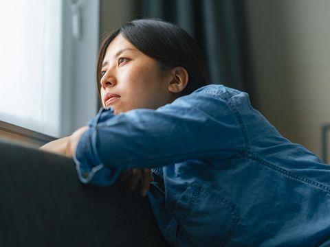 A young woman is sitting on a sofa by the window and looking outside through the window at home.