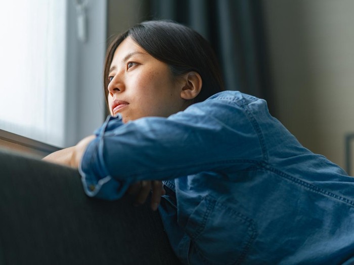 Ilustrasi Wanita Sedih dan Termenung A young woman is sitting on a sofa by the window and looking outside through the window at home.