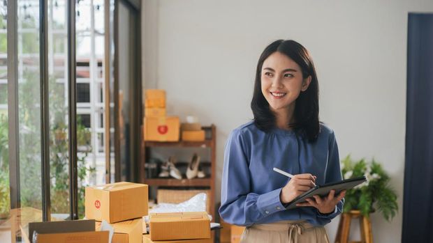 Portrait business Asian woman smile and use tablet checking information on parcel shipping box before send to customer. Entrepreneur small business working at home. SME business online marketing.