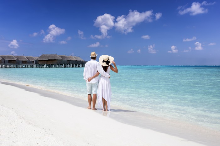 A hugging couple in white summer clothing stands on a tropical beach and enjoys the view to the turquoise sea and blue sky