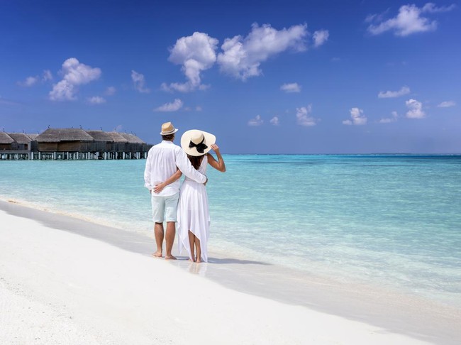A hugging couple in white summer clothing stands on a tropical beach and enjoys the view to the turquoise sea and blue sky