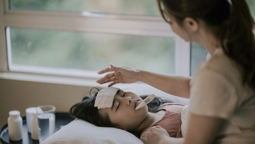 an asian chinese mother is taking care of her daughter on bed and measuring her body temperature with thermometer