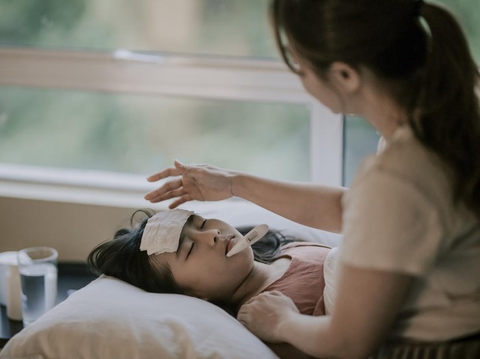 an asian chinese mother is taking care of her daughter on bed and measuring her body temperature with thermometer