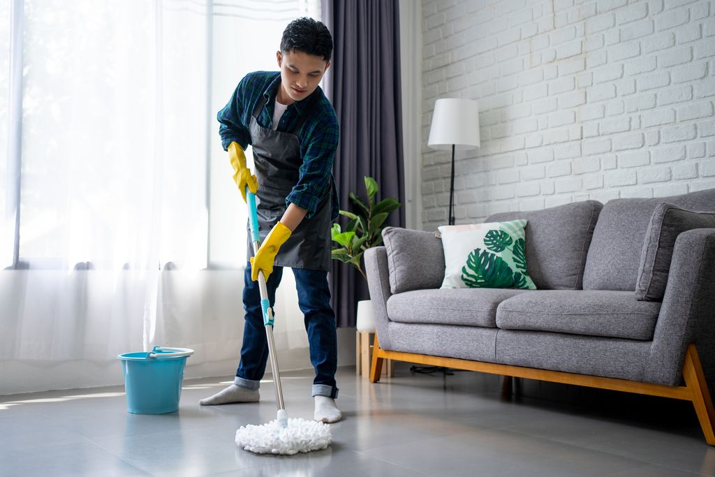 Handsome asian man wearing apron cleaning floor at home. Guy washing floor with mopping stick and bucket in living room.