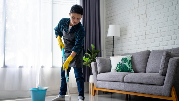 Handsome asian man wearing apron cleaning floor at home. Guy washing floor with mopping stick and bucket in living room.