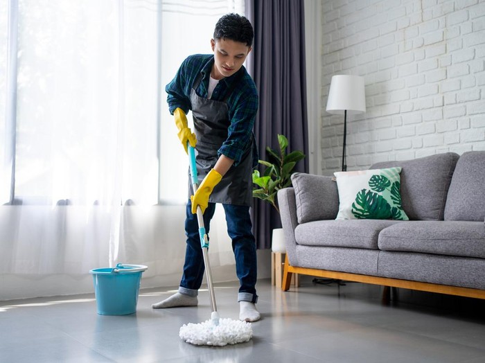 Handsome asian man wearing apron cleaning floor at home. Guy washing floor with mopping stick and bucket in living room.