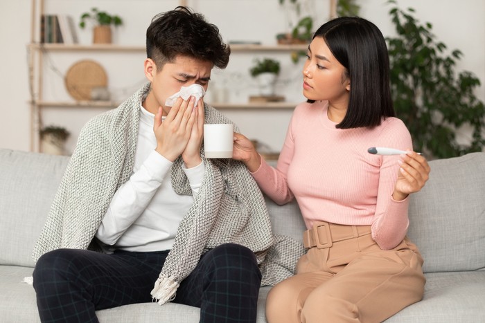 Chinese Wife Treating Ill Husband With Flu Giving Him Medicine Sitting On Sofa At Home. Seasonal Cold And Influenza Treatment Concept. Asian Girlfriend Takes Care Of Sick Boyfriend