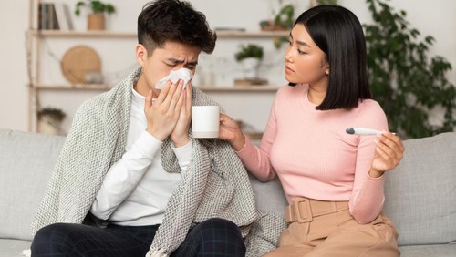 Chinese Wife Treating Ill Husband With Flu Giving Him Medicine Sitting On Sofa At Home. Seasonal Cold And Influenza Treatment Concept. Asian Girlfriend Takes Care Of Sick Boyfriend