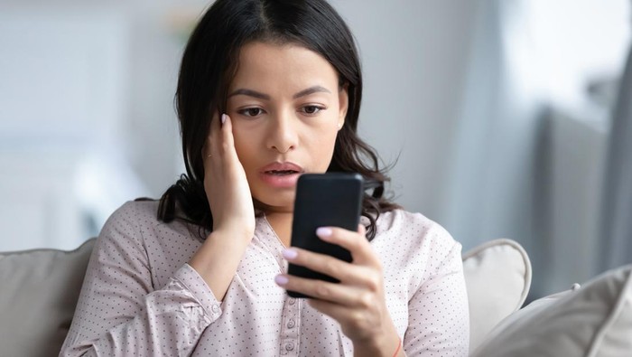 Upset African American young woman sit on couch look at smartphone screen shocked by negative news online, sad stunned biracial female read bad unpleasant notice or message on cellphone gadget