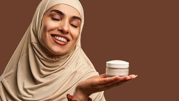 Portrait of beautiful modern muslim woman with natural make-up dressed in beige hijab smiling with closed eyes and holding cream in hand on brown background. Facial skin care, female beauty.