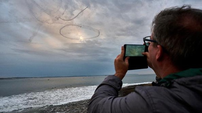 OEIRAS, PORTUGAL - OCTOBER 16: A spectator shoots video with his cellphone as Aerosparx Grob109b motor gliders perform aerobatics at the end of Oeiras Valley Air Show, an aerobatics festival integrated in the Portugal Air Summit'22, over Santo Amaro Beach on October 16, 2022 in Oeiras, Portugal. The festival included various aerobatics performances with airplanes from the Air Race Championship, a Supermarine Spitfire, and air shows by former world champions Ramón Alonso, Castor Fantoba, Jorge Macias, Luís Garção, Quijote, Jacobstars and The Raven Team. It also had an overflight of the Portuguese Air Force F16s and, at the end of the afternoon, Aerosparx team illuminating Santo Amaro water mirror with fireworks launched from the airplanes' wings. (Photo by Horacio Villalobos#Corbis/Corbis via Getty Images)