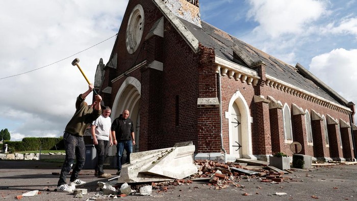 Tiga orang warga berdiri di depan gereja, salah satunya membawa palu untuk menghancurkan puing bangunan yang tergeletak di depan gereja tersebut.