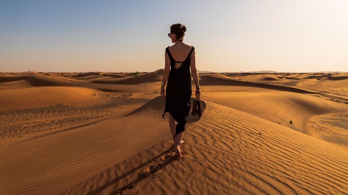 Lady in a black dress walking on the edge of the red dune in the middle of desert during windy spring weather
