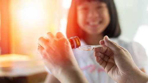 Happy Asia little child girl smiling want to take medicine form mother. Asian kid female waiting to eat drug. Hands of mom pouring cough syrup medicine into clear spoon to daughter.