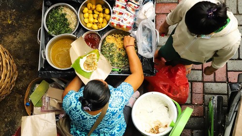 Ubud, Bali - January 11, 2020: Nasi Tepeng vendor in Ubud morning market. Nasi Tepeng is a local breakfast rice dish from Gianyar Regency in Bali. Here the lady vendor was preparing a portion as order came in. As this was a take-way order, so the dish was prepared in food paper that had been lined with banana leaf. Here seen the vendor scooping the vegetable dishes for the dish. A young lady was standing next to the tray; as she is the vendor assistant who takes order and handles payment. All the side dishes and condiments were laid on a very large metal tray which doubled as the serving counter. This stall is non-permanent and set up on a parking lot of Ubuds art market which in the morning used as morning market selling local food, including fresh fruits, vegetables and ready-to-eat meals.
