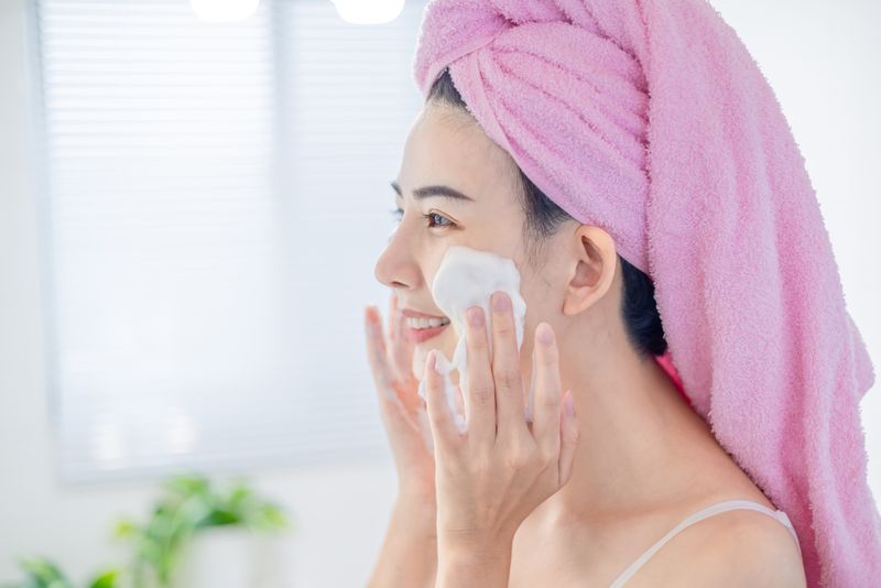 1372973043 asian beauty woman with towel on head is cleaning her face by facial cleanser in the bathroom