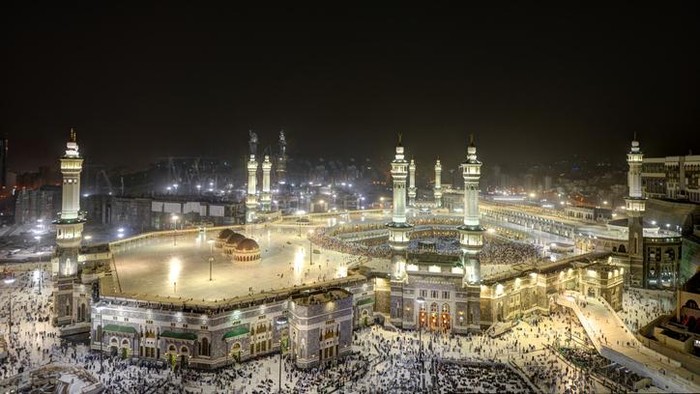 The ornate architecture around the Makkah Kaaba at night is alive with bright lighting that shows the people arriving for Hajj.  There are entrances to the structure on the ground level and along ramps up to the second story.  To the right, pilgrims encircle the black stone of Mecca.