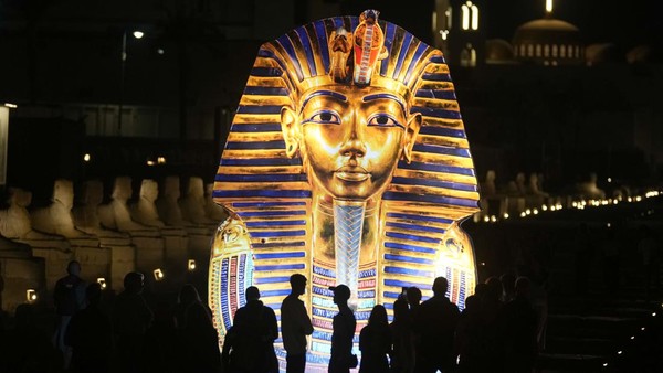 Turis berdiri di depan spanduk raksasa yang menunjukkan topeng emas Raja Tutankhamun di kuil Luxor di Luxor, Mesir, Jumat, 4 November 2022.