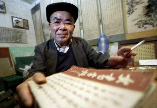 Zhou Shuoyi memegang salinan kamus pertama untuk bahasa Nushu yang diterbitkannya pada tahun 2003, saat ia berpose di rumahnya yang penuh dengan gulungan bahasa (latar belakang), di Jiangyong, ujung selatan China tengah. Provinsi Hunan. Pria tua itu adalah penjaga bahasa kuno yang penuh teka-teki, yang diyakini oleh para ahli bahasa Cina sebagai satu-satunya aksara di dunia yang secara eksklusif digunakan oleh wanita, yang diturunkan dari generasi sebelumnya, dan yang ia bayar dengan dua puluh satu tahun hidupnya. kehidupan ketika semangat komunis yang terlalu bersemangat mendidih menjadi perburuan penyihir dan kekerasan kolektif di tahun 1960-an oleh Mao Zedong. AFP PHOTO/Frederic J. BROWN (File Dok. FREDERIC BROWN / AFP)
