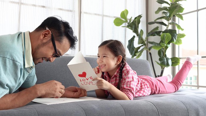 Happy Fathers Day, adorable cute little girl with young man dad lying on sofa in living room, making handmade greeting card for calibration. Happy family daughter and father spending time together.